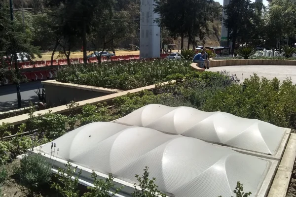 Vista amplia de una zona ajardinada urbana con domos de iluminación integrados en el suelo, cerca de edificios y un puente elevado.